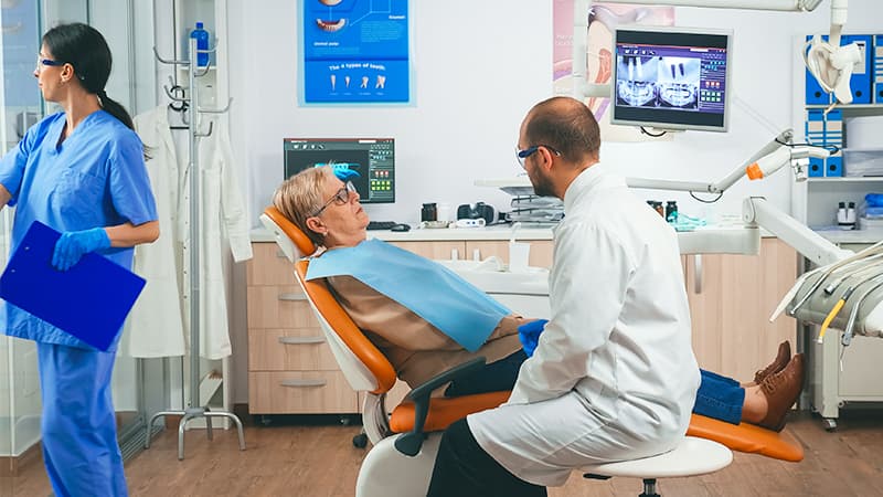 A dental specialist consults with a patient in the operatory while a coordinator works at the front desk station.