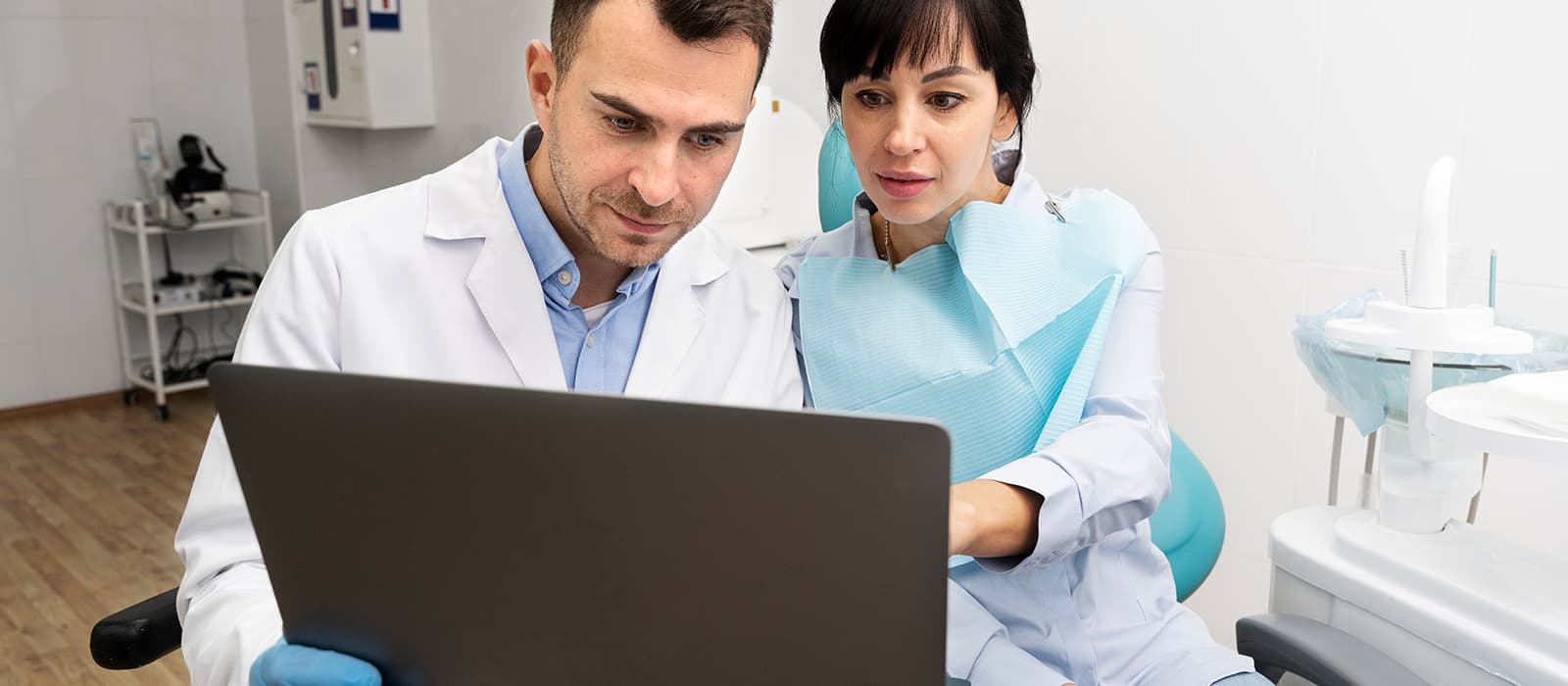 Two dental staff at a workstation review a referral dashboard on a wide monitor in a treatment room.