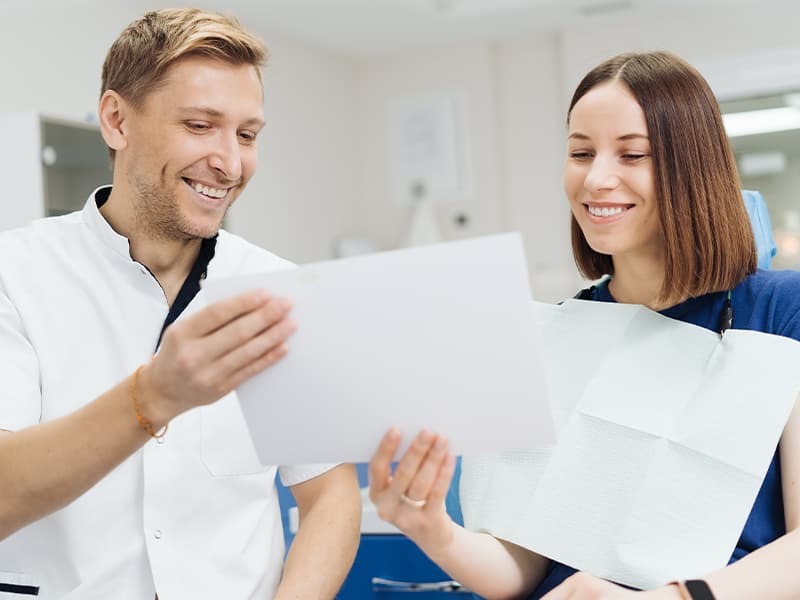 A dentist shows a treatment document to a smiling patient seated in the dental chair.
